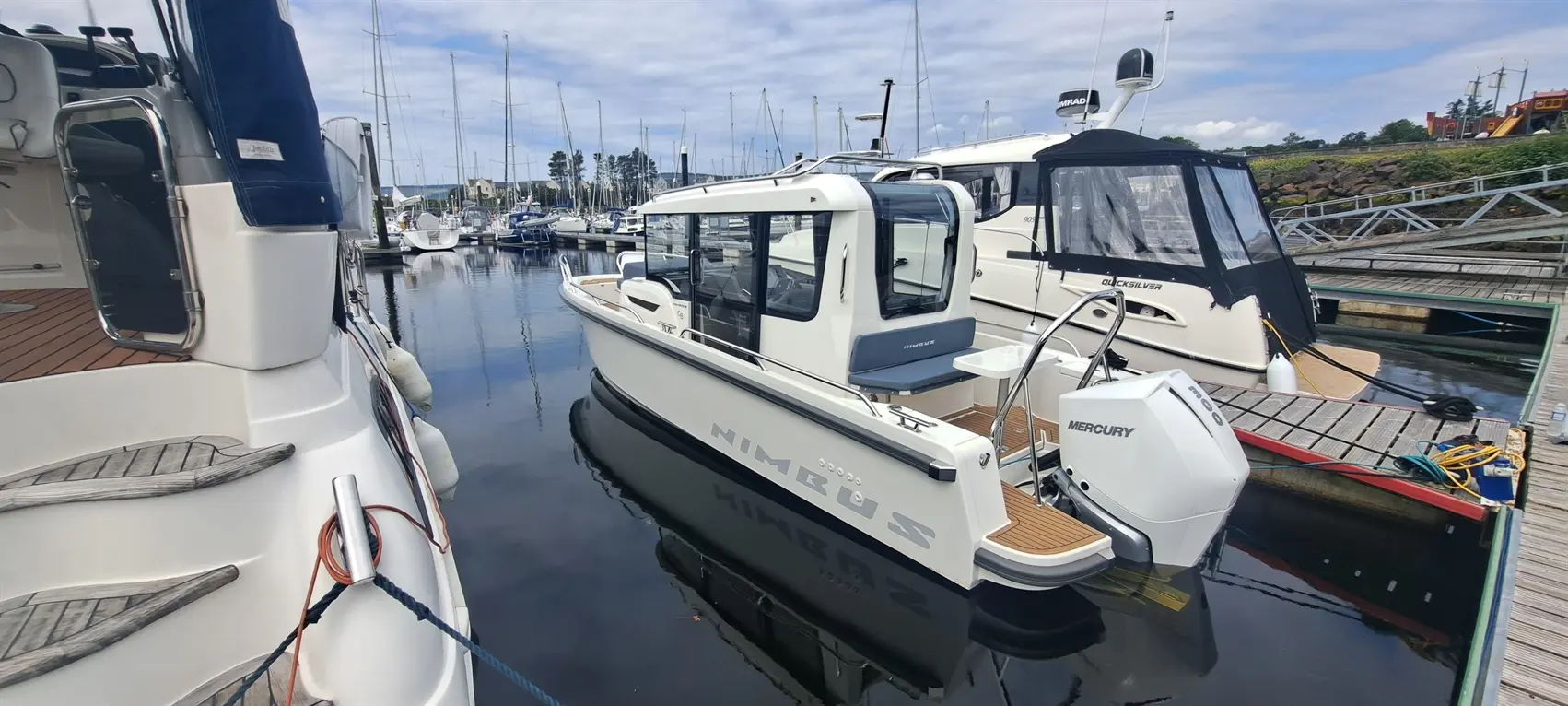 A Nimbus boat docked at a marina, next to another vessel, with sailing masts in the background and clear skies above.